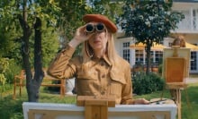 A blonde woman in a tan dress and an orange beret holds up a pair of binoculars while painting at an easel in a garden.