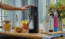 The SodaStream Sparking Water Maker resting on a kitchen counter surrounded by its accessories, while someone is touching its top.