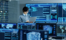 Someone holding up a computer while standing, surrounded by monitors displaying complex sets of data.