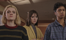 Three women stand in a hotel staring towards the camera with serious expressions.