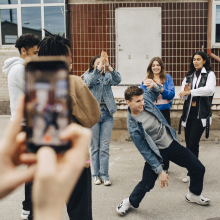 A person holds an iPhone and records a video of a young man dancing, surrounded by people clapping. 
