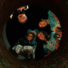 Four people look down a well, with the camera perspective below them inside the hole