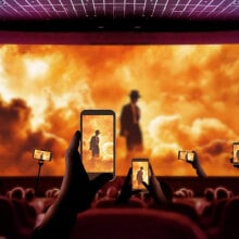 People using their phones to record a movie theater screening of "Oppenheimer"; the screen shows a thin silhouette of a man in a wide-brimmed hat, surrounding by fiery orange clouds. 