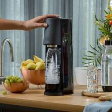 The SodaStream Sparking Water Maker resting on a kitchen counter surrounded by its accessories, while someone is touching its top.