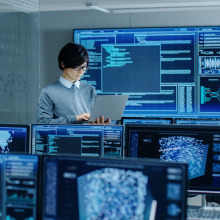 Someone holding up a computer while standing, surrounded by monitors displaying complex sets of data.