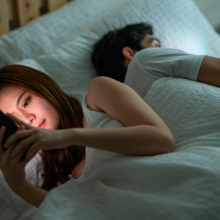 A young couple in bed, both facing opposite directions and staring at their phones. 