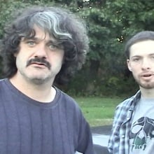 Two men stand outside outside, one is pointing at the camera.