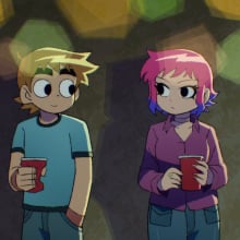 A man in a blue t-shirt and a woman with pink hair and pink clothes lean against the wall.