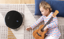 The Eufy BoostIQ RoboVac 11S on a carpet while a little girl sleeps next to it, holding a small guitar.