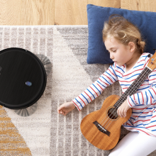 The Eufy BoostIQ RoboVac 11S on a carpet while a little girl sleeps next to it, holding a small guitar.