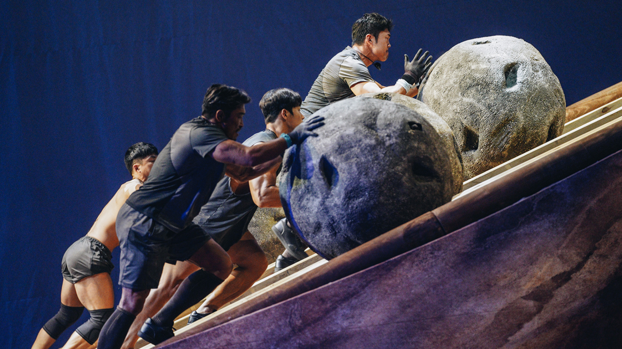 A group of men push boulders up an incline. 